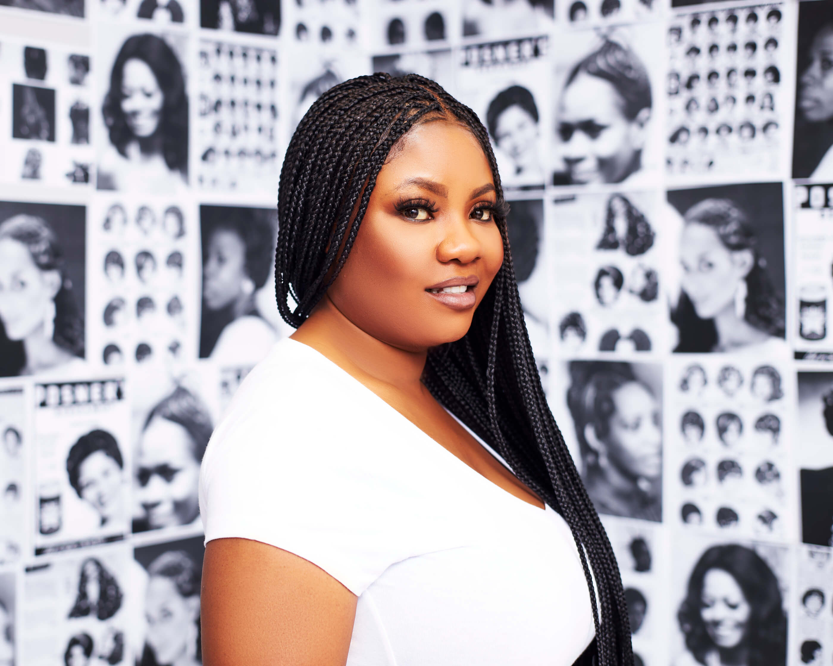 A woman with long braids posing in front of a picture-covered wall.